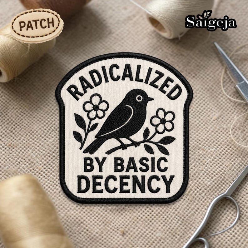 May include: Embroidered patch with the words "RADICALIZED BY BASIC DECENCY" and a black bird perched on a branch with flowers. The patch is cream-colored with black stitching. A small wooden patch that says "PATCH" is also visible.