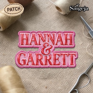 May include: A pink and red embroidered patch with the names "HANNAH & GARRETT" in a decorative font. The patch is on a beige fabric surface, with spools of thread and small scissors in the background.