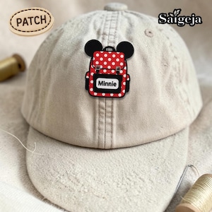 May include: A beige baseball cap featuring a red and white polka-dot backpack patch with black mouse ears. The patch includes the text Minnie. An oval tag labeled PATCH is in the upper left, and the brand name Saigeja is in the upper right.