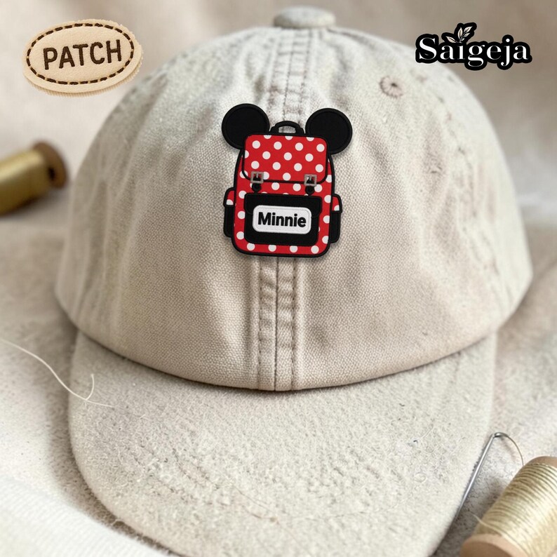 May include: A beige baseball cap featuring a red and white polka-dot backpack patch with black mouse ears. The patch includes the text Minnie. An oval tag labeled PATCH is in the upper left, and the brand name Saigeja is in the upper right.