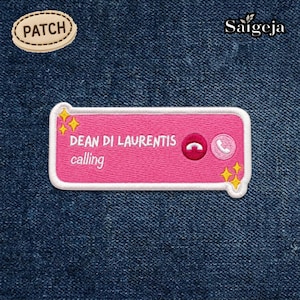 Puede incluir: Parche bordado con una pantalla de teléfono rosa con el texto "DEAN DI LAURENTIS calling". El parche incluye un icono de teléfono, un icono de llamada y detalles de estrellas amarillas. También es visible un parche más pequeño con la palabra "PATCH".