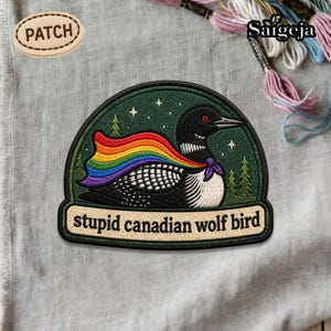 May include: Embroidered patch featuring a black and white bird with a rainbow cape, set against a dark green background with stars and trees. The text "stupid canadian wolf bird" is at the bottom. A smaller patch with the word "PATCH" is at the top.