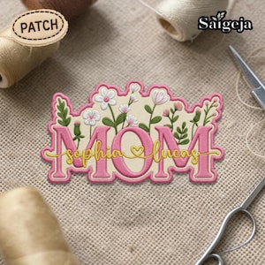 May include: Embroidered patch with the word "MOM" in pink, with "Sophia & Lucas" in cursive script. The patch features floral and leaf accents in white and green. The word "PATCH" is on a small wooden tag.