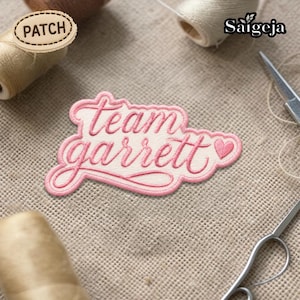 Puede incluir: Parche bordado rosa y crema con las palabras "Team Garrett" en cursiva y un corazón. El parche está sobre una superficie texturizada, con carretes de hilo y tijeras en el fondo. Ideal para personalizar ropa.