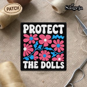 以下が含まれることがあります： 白いブロック文字で「PROTECT THE DOLLS」と書かれた黒い正方形のパッチ。白い中心と青い茎とアクセントが付いたピンクの花がパッチを飾っています。パッチは縫製用品に囲まれています。