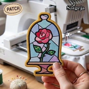 Peut inclure: Écusson brodé représentant une rose rose dans une cloche en vitrail, avec une bordure dorée. Le patch est tenu par une main. Le mot "PATCH" est sur un patch ovale en bois. Une machine à broder est en arrière-plan.