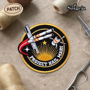Puede incluir: Parche bordado con borde negro. El diseño presenta un cohete, una estrella y el texto "PROJECT HAIL MARY". El parche utiliza colores blanco, naranja y amarillo. Ideal para ropa o accesorios.