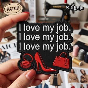 May include: Black embroidered patch with the text "I love my job" repeated three times. The patch features red high heels, a compact, and a handbag. The patch is held up by a person's hand.