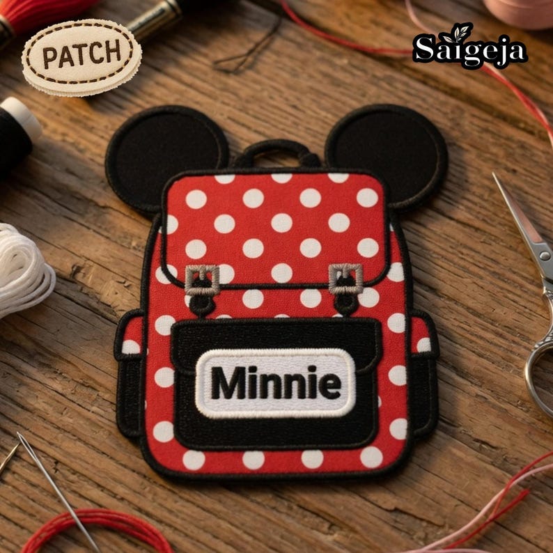 May include: A red and white polka dot embroidered patch shaped like a backpack with black mouse ears at the top. The patch features a black front pocket with a white rectangular label containing the black text Minnie and two silver buckles.