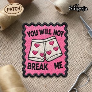 Op de afbeelding: Een roze geborduurde patch met de tekst "YOU WILL NOT BREAK ME" en een illustratie van witte shorts met roze harten. De patch heeft een zwarte rand en een decoratieve rand. Het woord "PATCH" staat op een houten label.
