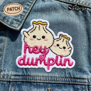 Puede incluir: Parche bordado con dos personajes de dumpling sonrientes con mejillas rosadas y detalles amarillos. El parche tiene el texto "hey dumplin" en letras cursivas rosas. El parche está sobre una chaqueta vaquera.
