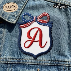 Op de afbeelding: Geborduurde patch met een wit schild en een rode letter "A" in script. Het schild heeft een marineblauwe geschulpte rand en een rood-wit-blauwe strik. De patch is bevestigd op een spijkerjasje.