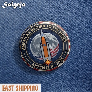 May include: A round button with the text "AMERICA'S RETURN TO THE MOON" and "ARTEMIS II 2026". The button features a rocket, the moon, and the American flag in a blue, orange, and white color scheme.