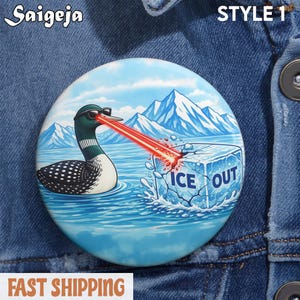 May include: A round button with a cartoon loon wearing sunglasses, shooting a red laser beam from its eyes, melting an ice cube that says "ICE OUT". The background features a lake and mountains. The button has the text "STYLE 1".