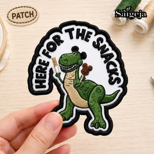 Pode incluir: Um emblema bordado apresentando um dinossauro verde com barriga bege, usando orelhas de rato pretas. O dinossauro segura um churro e um doce de chocolate. O texto HERE FOR THE SNACKS está bordado em preto sobre um fundo branco.