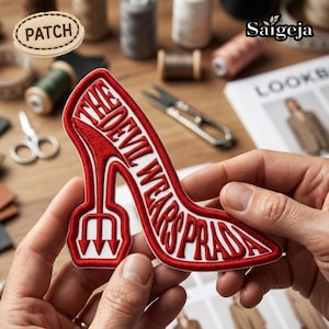 May include: A red and white embroidered patch in the shape of a high-heeled shoe. The text "THE DEVIL WEARS PRADA" is inside the shoe. The patch is being held by a person.