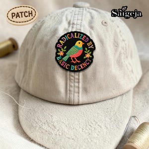 Pu&ograve; includere: Un cappellino da baseball beige con una toppa colorata. La toppa presenta un disegno di uccello con le parole "RADICALIZED BY BASIC DECENCY" in un motivo circolare. &Egrave; visibile anche una toppa separata con la parola "PATCH".