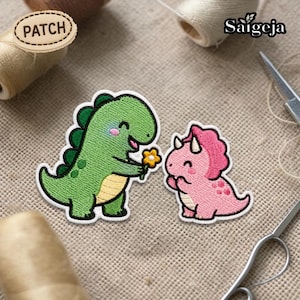 May include: Embroidered patch set featuring two cartoon dinosaurs. One green dinosaur offers a yellow flower to a pink dinosaur. The patches have white borders and are on a tan fabric background. The word "PATCH" is on a wooden label.