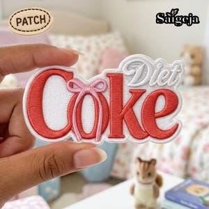May include: An embroidered patch featuring the text Diet Coke in red and white thread. The letter O is stylized with a light pink ribbon tied in a bow. The patch has a white border and a white background, with the word PATCH printed in a small oval.