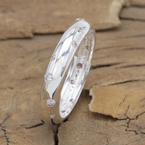 May include: A polished silver-toned ring featuring a smooth, rounded band with multiple small, clear, circular stones embedded at irregular intervals along the outer surface, creating a scattered, flush-set appearance.