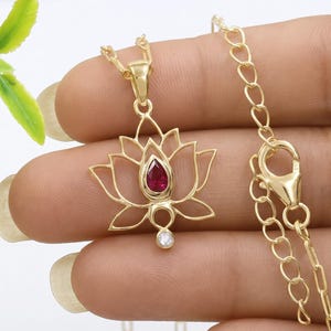 May include: A gold-toned necklace featuring a lotus flower pendant. The pendant is centered with a pear-shaped red gemstone and a small, round, clear stone at the base. The chain consists of oval links and includes a lobster claw clasp closure.