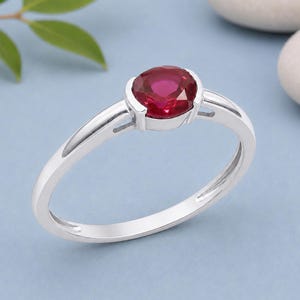 May include: A silver ring featuring a vibrant, oval-cut ruby gemstone. The ring has a simple, elegant design with a polished band and a bezel setting that securely holds the red gemstone. The ring is displayed on a light blue surface.