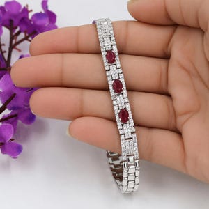Peut inclure: Un bracelet argenté orné de trois pierres précieuses ovales rouge foncé et de nombreuses petites pierres claires. Le bracelet est rectangulaire et tenu dans une main.