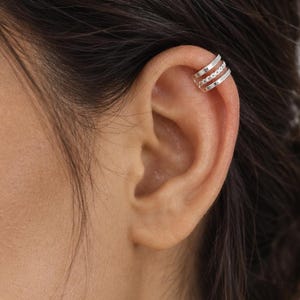 May include: A silver-toned ear cuff featuring three distinct bands. The top and bottom bands are smooth and polished, while the middle band displays a textured, beaded design. This piece of jewellery is worn on the upper cartilage of the ear.