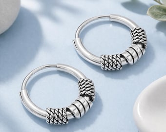 May include: A pair of silver hoop earrings featuring a smooth circular band accented with two distinct textured segments. Each earring includes a braided rope-like section and a smooth, slightly curved metal band detail in a polished finish.