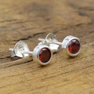 May include: A pair of silver stud earrings with round, deep red gemstones. The earrings are set in a simple bezel setting, showcasing the vibrant color of the stones. The earrings are displayed on a wooden surface.