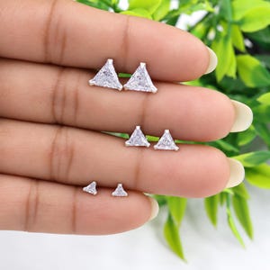 May include: Three pairs of triangle-shaped stud earrings in varying sizes, each featuring a clear, faceted stone set in a silver-toned metal base. The earrings are arranged in descending order of size from top to bottom on a flat surface.