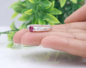 May include: A silver ring featuring a single round, deep red gemstone set in the centre. The band is smooth and polished, with a simple, clean design that highlights the vibrant colour of the stone against the metallic silver finish.