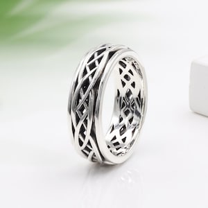 May include: A silver ring with a detailed, intricate, woven design. The ring is wide and has a smooth, polished finish.
