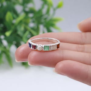 Op de afbeelding: Een zilveren ring met een rij kleurrijke edelstenen en een centrale parel. De edelstenen zijn vierkant geslepen en bevatten blauwe, rode, groene en oranje tinten. De ring wordt door een hand vastgehouden, wat het ontwerp en het vakmanschap benadrukt.