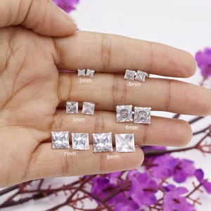May include: A selection of square-cut stud earrings in various sizes, from 3mm to 8mm. The earrings are shown on a hand, highlighting their clear, sparkling appearance. Sizes are labelled in millimetres.