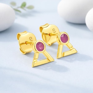 以下が含まれることがあります： 三角形のデザインと丸いピンク色の宝石が付いた、金色のスタッドピアス。 ピアスには、安全な着用を可能にするポストと裏地が付いています。 ピアスは水色の表面に表示されています。