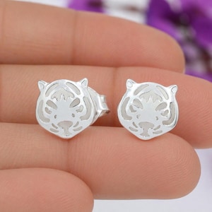 May include: A pair of silver-colored stud earrings. Each earring is shaped like a tiger's head with cut-out details. The earrings are held in a person's hand, with a blurred background.