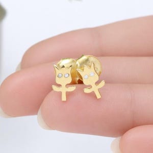 May include: A pair of gold-colored stud earrings. Each earring features a stylized flower design with a crown and two small, clear gemstones. The earrings are held between fingers, showcasing their size and design.