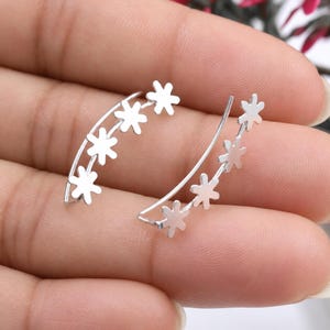 May include: A pair of silver-toned ear climbers, each featuring a curved wire design adorned with four small, six-petaled flower shapes arranged in a row along the length of the metal wire, creating a delicate and symmetrical appearance.