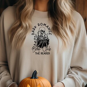May include: Beige sweatshirt with a black graphic of a skeleton reading a book, surrounded by the words "Dark Romance Book Club The Reader." A carved orange pumpkin is held in front of the sweatshirt.