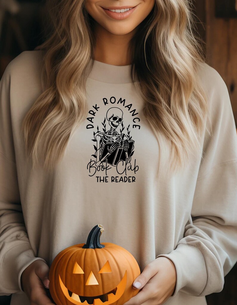 May include: Beige sweatshirt with a black graphic of a skeleton reading a book, surrounded by the words "Dark Romance Book Club The Reader." A carved orange pumpkin is held in front of the sweatshirt.