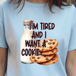 Puede incluir: Una camiseta azul claro con el texto "I'm tired and I want a cookie" en texto marrón. Una botella de vidrio de leche y una pila de galletas con chispas de chocolate se muestran junto al texto.