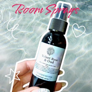 May include: A glass bottle of room spray with a black cap and label. The label is white with black text that reads "Velvet Rose & Oud" and "Room Spray Care Instructions". The bottle is sitting on a hand with a star and heart drawn around it.