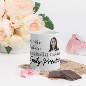 May include: White ceramic mug featuring the text "Single, Taken, Mentally dating Emily Prentiss" and a portrait of a woman. The mug is set amongst flowers, sweets, and chocolate.