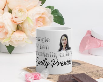 May include: White ceramic mug with the text "Single, Taken, Mentally dating Emily Prentiss" and a portrait of a woman. The mug is surrounded by flowers, candy, and chocolate.