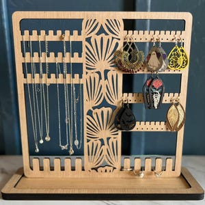 Peut inclure: Un présentoir à bijoux en bois avec plusieurs fentes pour colliers et boucles d'oreilles. Le présentoir a un motif découpé décoratif et un plateau à la base. Divers colliers et boucles d'oreilles, dont des pièces dorées et noires, sont exposés.