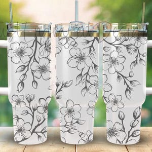 May include: Three stainless steel tumblers with a white background and a black floral design. The tumbler on the left has a handle.