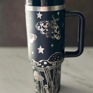 K&ouml;nnte beinhalten: Schwarze Reise-Thermoskanne aus Edelstahl mit einem silbernen Design aus Pilzen, Sternen und einem Nachtfalter. Der Nachtfalter hat einen Halbmond und Sterne auf seinen Fl&uuml;geln.