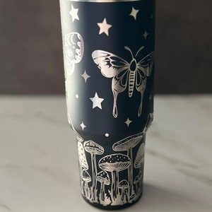 K&ouml;nnte beinhalten: Ein schwarzer Edelstahlbecher mit einem silbernen Design aus Pilzen, Schmetterlingen und Sternen.