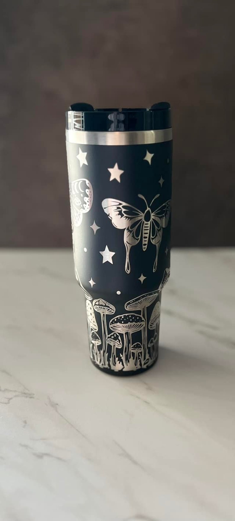 K&ouml;nnte beinhalten: Ein schwarzer Edelstahlbecher mit einem silbernen Design aus Pilzen, Schmetterlingen und Sternen.